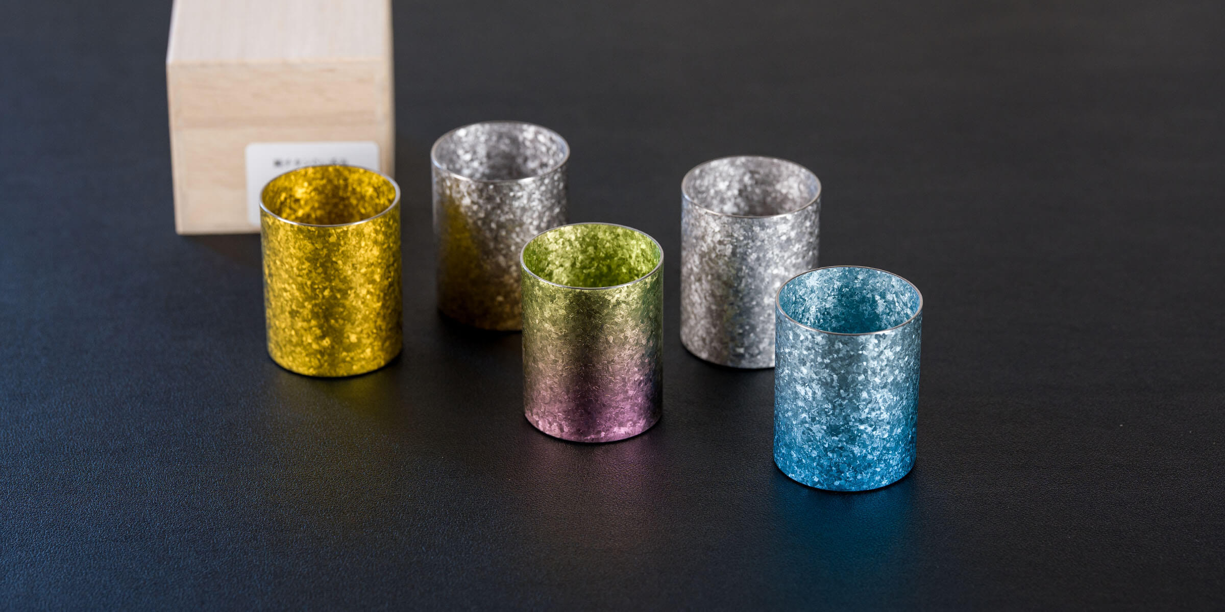 Group of colorful HORIE Titanium sake cups arranged on a dark surface, highlighting their brilliant metallic luster.